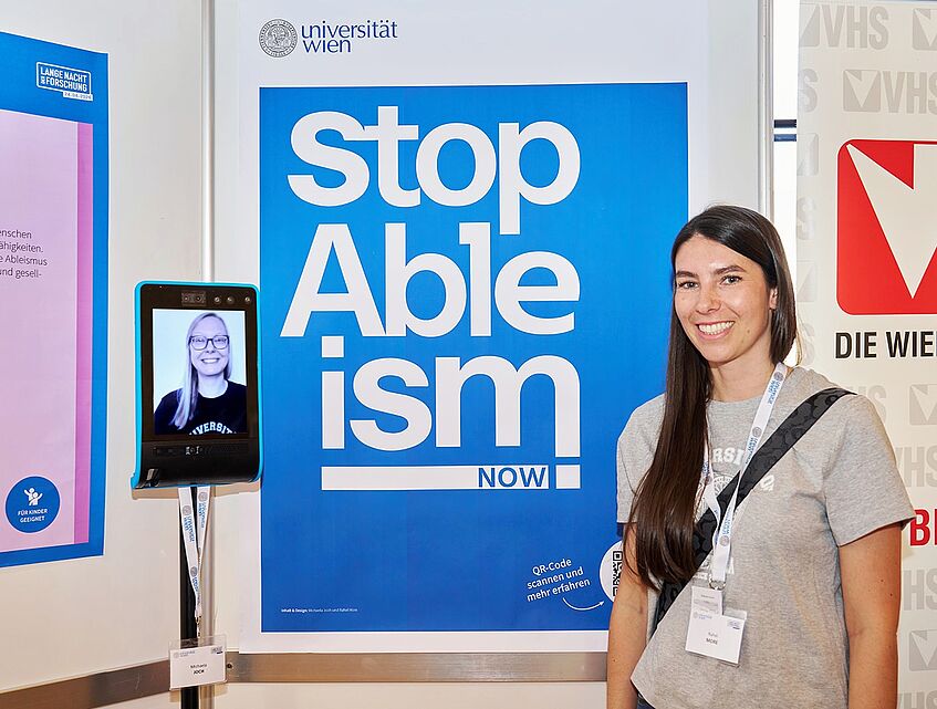 LNF26 Rahel More steht rechts von einem Plakat auf dem Stop Ableism Now! steht. Links davon steht ein Telepräsenzroboter. Das ist wie ein Tablet auf einer Stange mit zwei Rollen untendran zur Fortbewegung. Auf dem Bildschirm sieht man Michaela Joch.